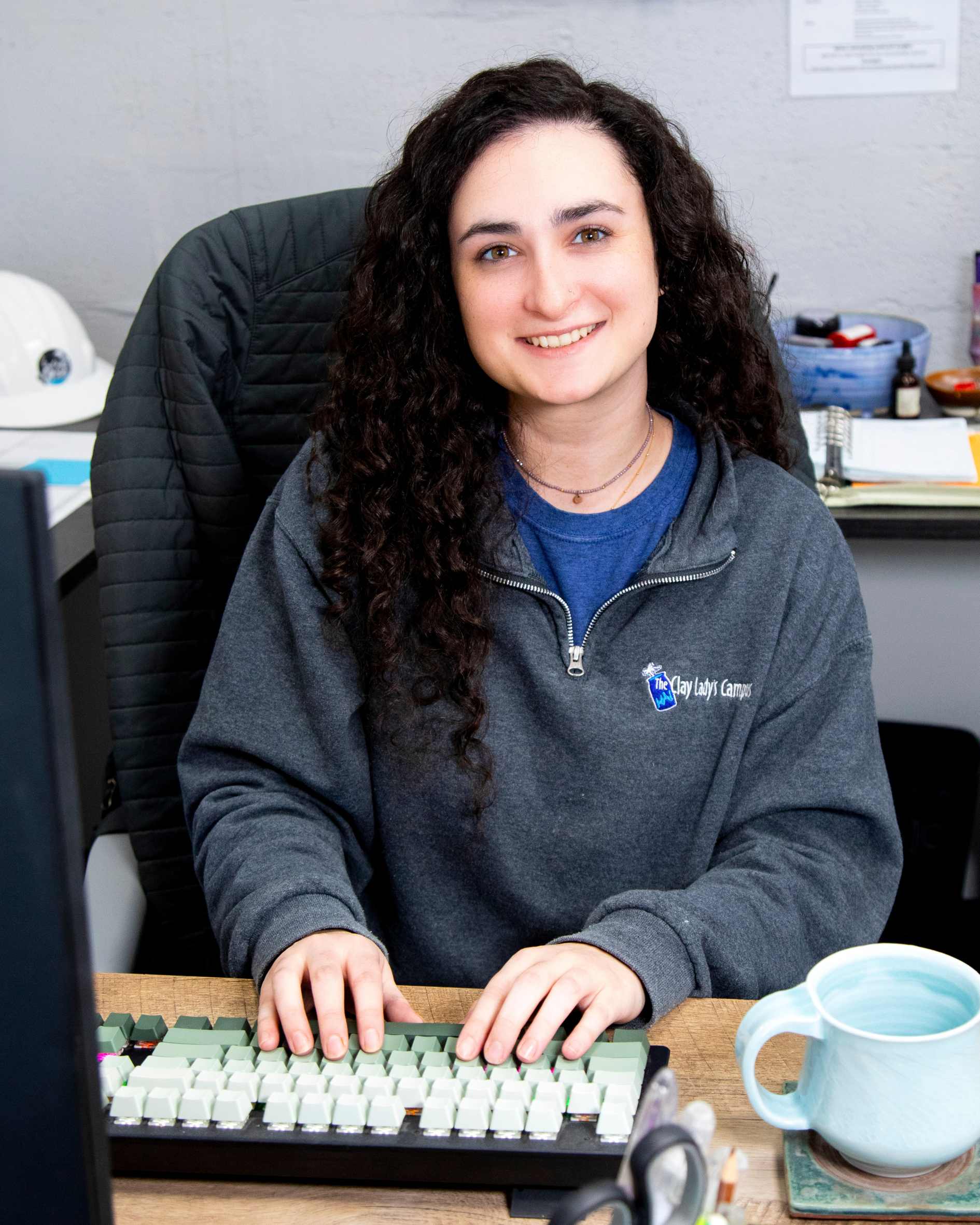 Celeste Vangeli, with curly hair wearing a gray Clay Lady hoodie, in an office setup.
